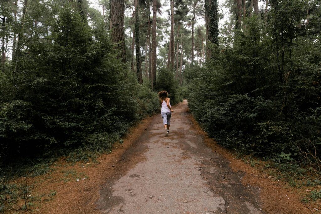 A woman runs on a forest trail surrounded by lush greenery during the day.